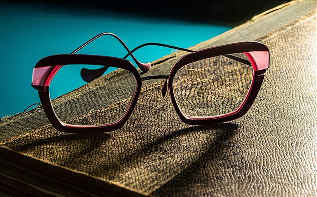 Eine Brille mit einem modernen, pinken Rahmen liegt auf einem alten Buch. Der Hintergrund ist in einem sanften Blau.