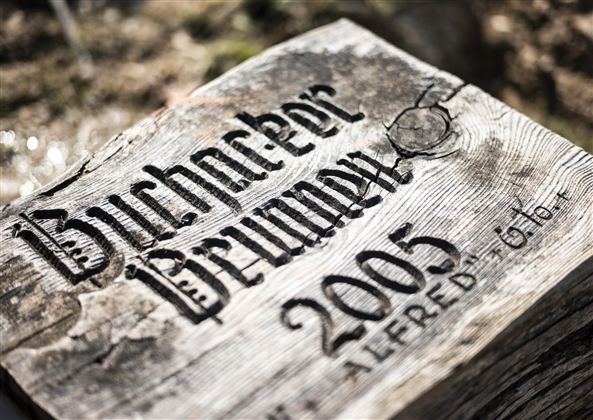 Een houten bord met de inscriptie "Buchacker Brünnlein 2005". De letters zijn in een artful lettertype gegraveerd.