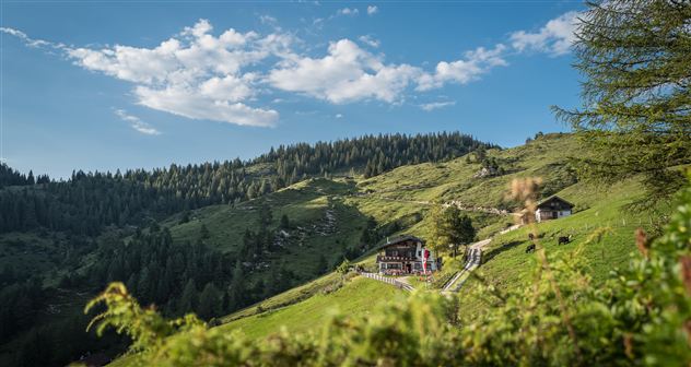 Een schilderachtig herberg in een bergachtig landschap. Twee wandelaars lopen voorbij op een smal pad.