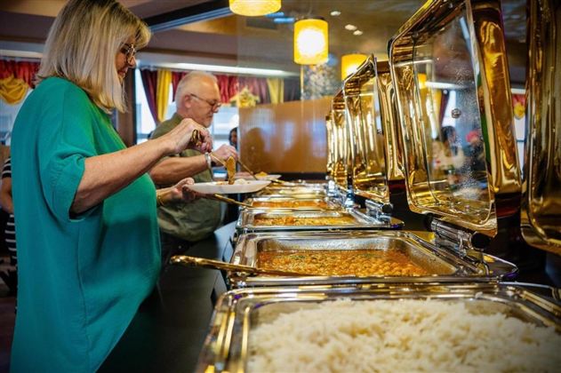 A woman and a man are serving themselves at a buffet with various dishes. In the foreground, rice is visible, while the selection of foods is presented behind glass bowls.