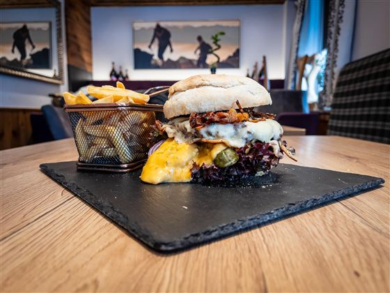 Ein saftiger Burger mit Käse und frischem Gemüse liegt auf einem schwarzen Servierteller. Dazu gibt es knusprige Pommes frites in einem kleinen Korb.