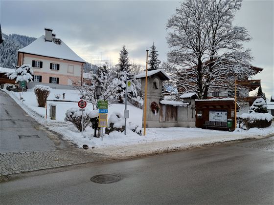 Eine verschneite Straße in einem ruhigen Dorf. Im Hintergrund sind Häuser und verschneite Bäume zu sehen.