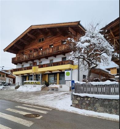 Ein traditionelles Gebäude mit Holzfassade und einem weißen Erdgeschoss. Draußen liegt Schnee, und es gibt eine Bank mit dem Schriftzug „Eisenbank“.