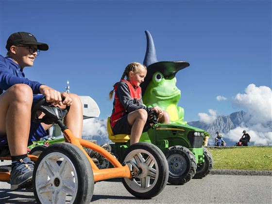 Two children are riding small vehicles on a sunny meadow. In the background, a large green figure with a wizard hat can be seen.