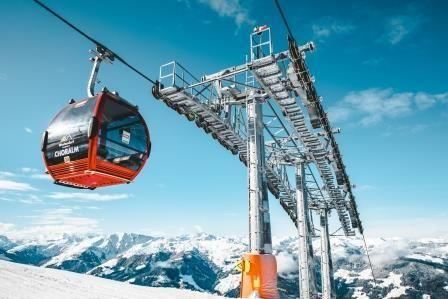 A cable car glides over a snowy mountain landscape. In the background, impressive mountains and a clear blue sky can be seen.