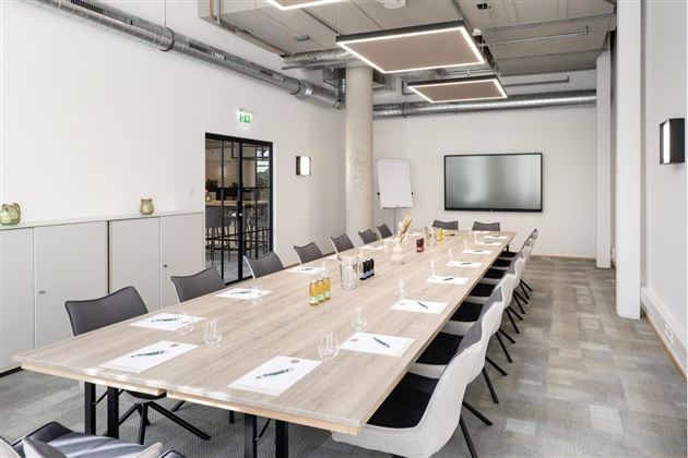 A modern meeting room with a long table and chairs.  
On the wall hangs a large screen, and there are various drinks and writing materials on the table.