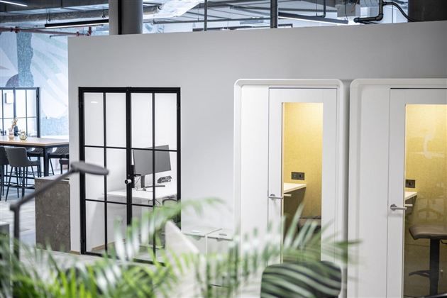 A modern office with glass meeting rooms and closed phone booths. In the foreground, there are green plants that create a pleasant atmosphere.