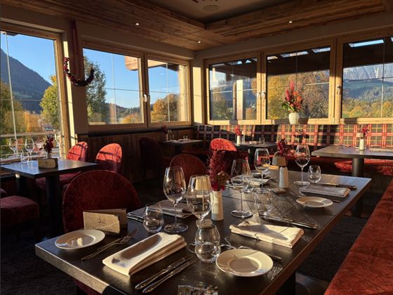 An elegant restaurant with wooden decor and red chairs. The table is festively laid with glasses and dishes, and large windows offer a view of the landscape.
