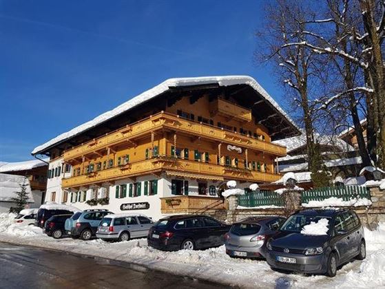 Een traditioneel Alpijns gebouw met een houten balkon en groene luiken. De omgeving is bedekt met sneeuw en er staan verschillende auto’s op de weg.