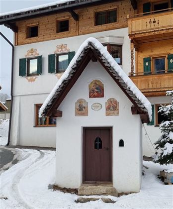 Eine kleine Kapelle mit einem schneebedeckten Dach und kunstvollen Fresken. Im Hintergrund sind traditionelle Holzhäuser zu sehen.
