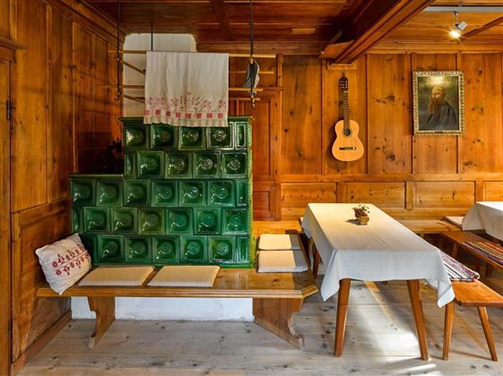 Ein gemütlicher Raum mit Holzwänden und einem traditionellen grünen Ofen. Auf den Tischen liegen weiße Tischdecken und eine Gitarre hängt an der Wand.