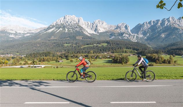 Two cyclists ride along a road on a vast green meadow. In the background, impressive mountains and a clear blue sky can be seen.
