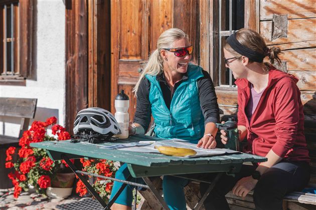 Two women are sitting at a table outdoors and are chatting. On the table, there is a bicycle helmet, a water bottle, and a banana.