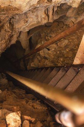 A steep metal staircase leads into a dark cave. The floor is covered with stones and earth.
