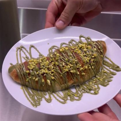 A freshly baked croissant on a plate, garnished with green drizzle and chopped pistachios. The dessert looks appetizing and tempting.
