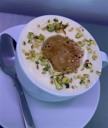 A cup with creamy drink, garnished with chopped pistachios. Next to the cup, there is a spoon on a saucer.
