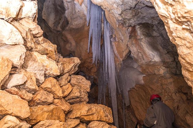 A cave with large rocks and stalagmites. A person with a red helmet is exploring the room.