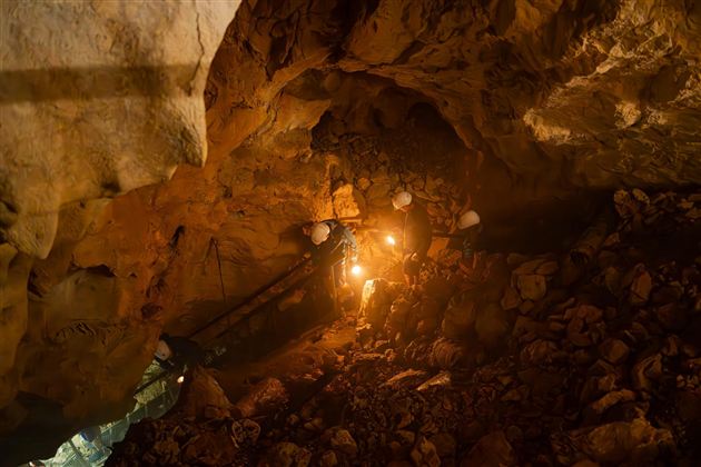 A gloomy cave chamber with rocks and a floor made of stones. Two people with helmets are holding flashlights and exploring the surroundings.