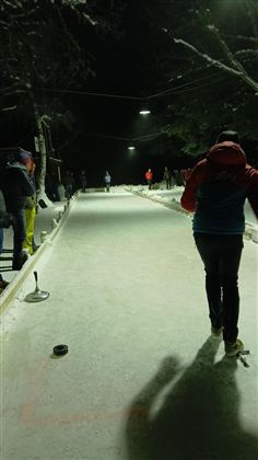 Eine verschneite Eisbahn bei Nacht mit beleuchteten Bereichen. Einige Personen sind beim Spielen aktiv.