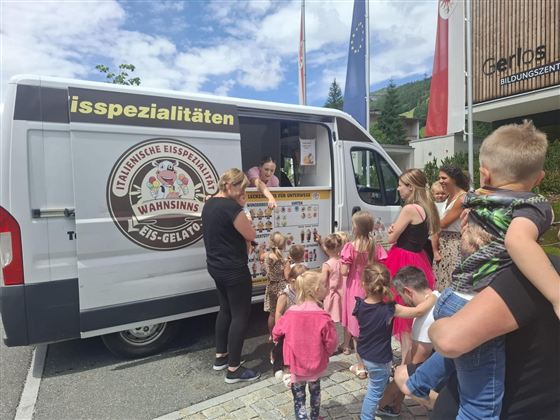 An ice cream truck with various ice cream specialties is parked on the street. Children and adults are standing in line to buy ice cream.