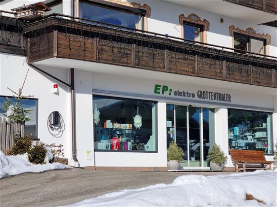Ein Elektrogeschäft mit großen Fenstern sorgt für einen Blick auf verschiedene Produkte. Vor dem Geschäft liegt Schnee, und die Umgebung ist typisch für eine Berglandschaft.