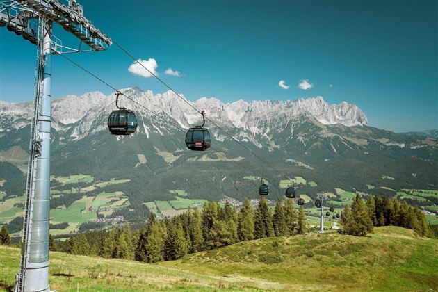 A cable car travels over a green landscape with mountains in the background. The sky is clear and blue.