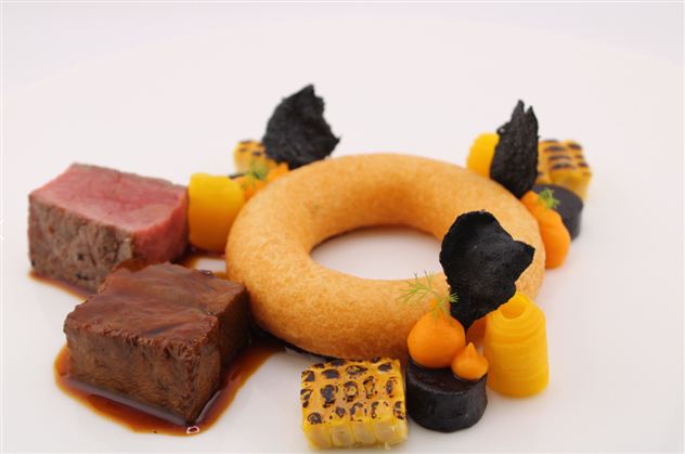 An artistically arranged dish with various cuts of meat, a round, donut-like side, and colorful vegetables. The presentation emphasizes the different textures and colors.