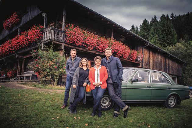 Eine Gruppe von vier Personen steht vor einem Holzhaus mit blühenden Balkonpflanzen. Ein grünes Auto befindet sich neben ihnen auf einer Wiese.