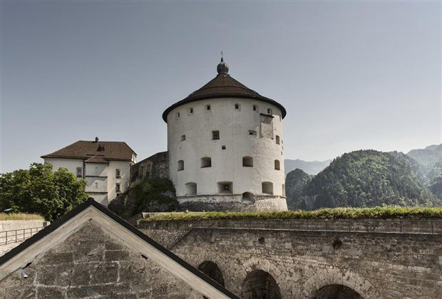 An old fortress with a round tower and a picturesque view of the mountains. The landscape is green and mountainous, creating a peaceful atmosphere.