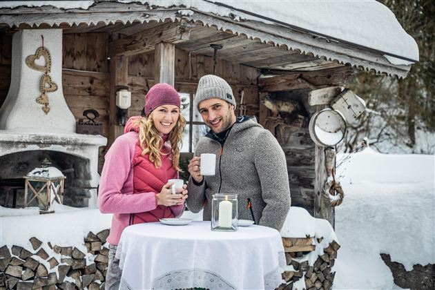 Ein Paar steht lächelnd an einem Tisch im Schnee und hält Tassen in der Hand. Im Hintergrund ist eine rustikale Hütte und gestapeltes Holz zu sehen.