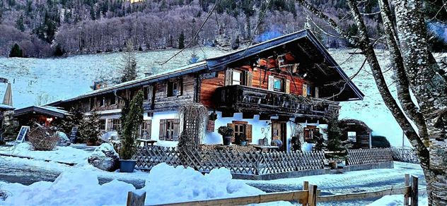 Ein traditionelles Chalet im Schnee, umgeben von Bäumen und Bergen. Die Fassade ist aus Holz mit gemütlichen Balkonen und Fensterläden.