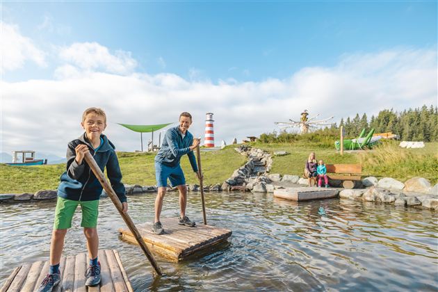 Two children are standing on a wooden platform in the water and having fun. In the background, there are green meadows and a lighthouse.