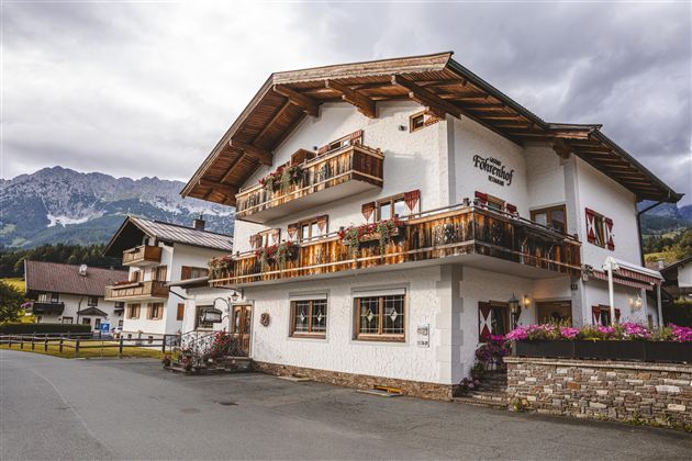 Een charmant, tweelaags huis met houten balkon en bloemen. Op de achtergrond zijn bergen en een rustige straat te zien.
