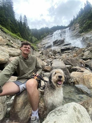 A young man is sitting on stones next to a dog and smiling. In the background, there is a waterfall and wooded hills visible.
