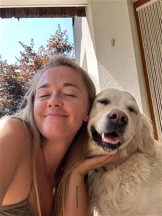 A woman smiles happily with a dog beside her.  
They enjoy a sunny day outdoors.