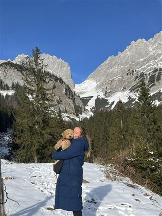 A woman holds a dog in her arms and smiles in nature. In the background, tall mountains and a clear blue sky can be seen.