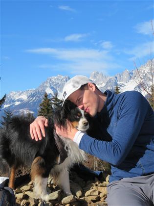 A man hugs his dog in the mountains. In the background, snow-covered mountains and a blue sky can be seen.