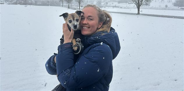 A smiling woman is holding a small dog in her arms. In the background, there is snow and it is lightly snowing.