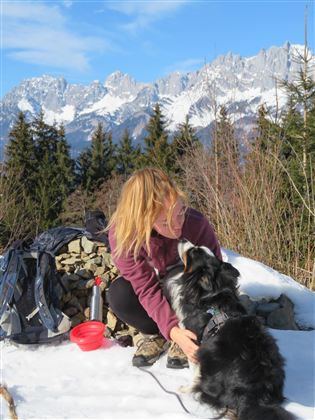 A man is sitting on a path and petting his dog. In the background, there are high mountains and a green valley.
