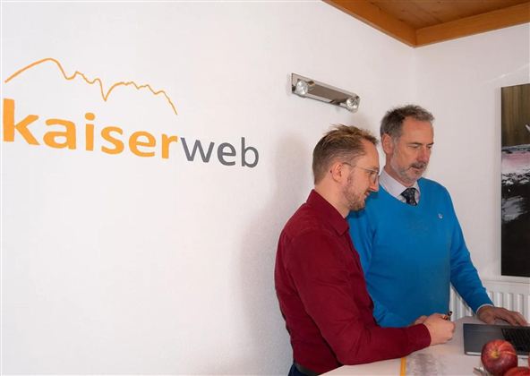 Two men are standing at a table and looking at a laptop. In the background, the logo of kaiserweb can be seen.