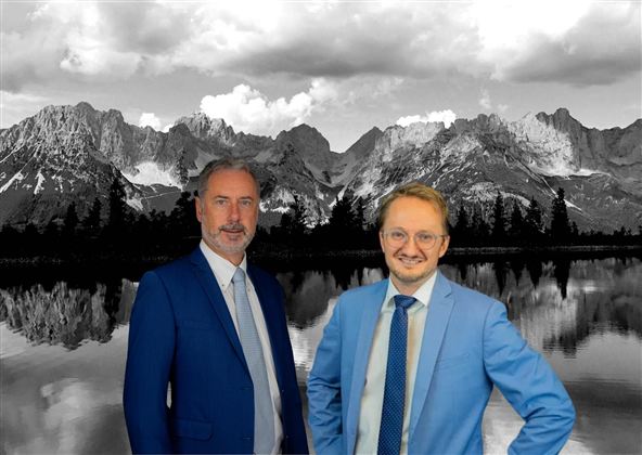 Two men in suits are standing in front of an impressive mountain landscape. The background shows the mountains and a calm lake, while the sky is cloudy.