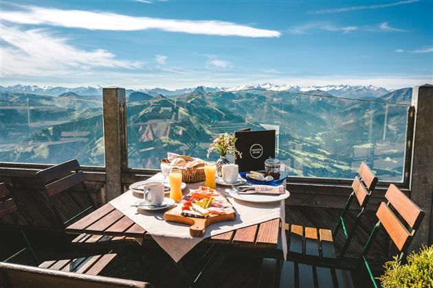 Een tafel op een terras met uitzicht op de bergen. Op de tafel staan ontbijgerechten, koffie en een sapbar.