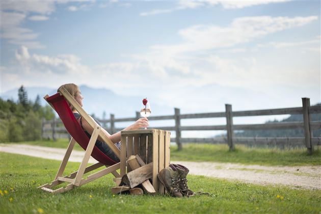 Een persoon zit ontspannen in een ligstoel op een grasveld. Op de achtergrond zijn bergen en een blauwe lucht te zien.
