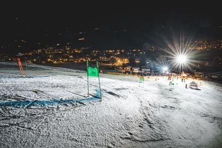 Eine beleuchtete Skipiste bei Nacht mit grünen Toren. Im Hintergrund sind Lichtpunkte von einem Dorf zu sehen.