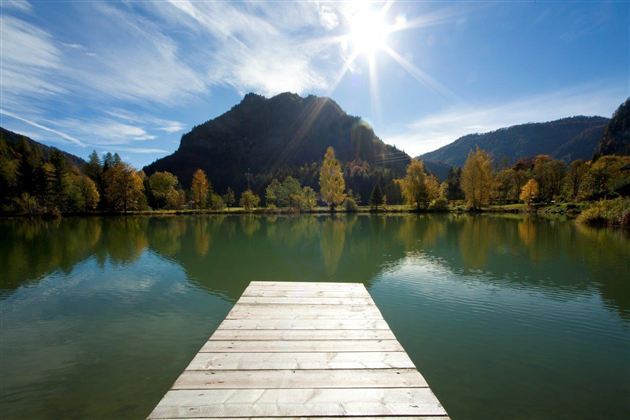 A quiet lake with a wooden pier and colorful trees in the background. The sun shines in the clear sky and reflects in the water.