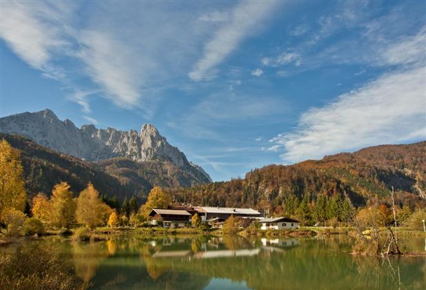 An idyllic landscape with a clear lake and colorful autumn foliage. In the background, majestic mountains and a blue sky can be seen.