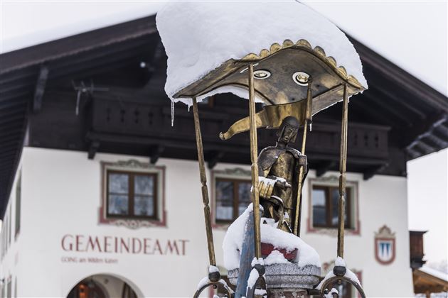 Ein Brunnen mit Schnee auf dem Dach und einer Statue im Vordergrund. Im Hintergrund steht ein traditionelles Gebäude mit der Aufschrift "Gemeindeamt".