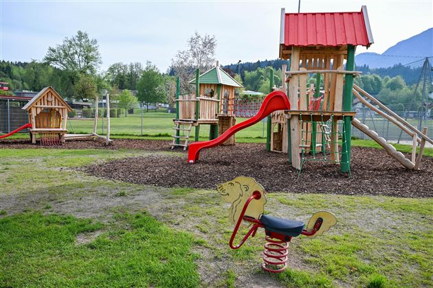 A playground landscape with wooden play equipment and a slide. In the foreground, there is a seesaw shaped like a horse.
