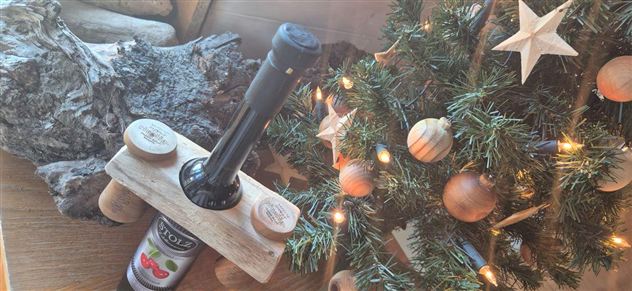 A wooden stand with a bottle and a corkscrew stands next to a decorated Christmas tree. In the background, natural elements such as wood and decorated baubles can be seen.