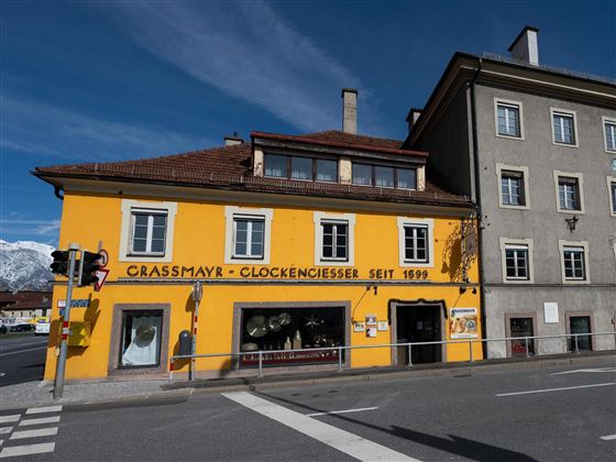 Ein gelbes Gebäude mit dem Schriftzug "Grassmayr - Glockengießerei seit 1599" steht an einer Straßenecke. Daneben befindet sich ein graues Haus und im Hintergrund sind Berge zu sehen.